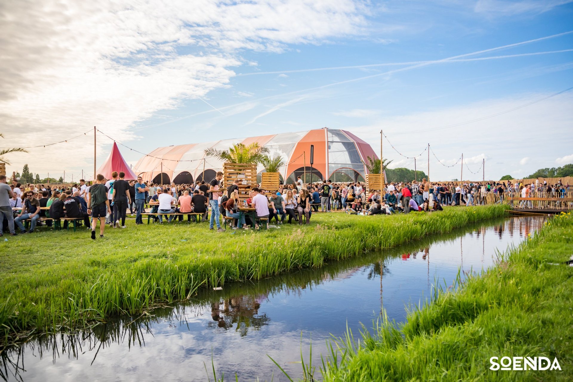 Een grote groep mensen op een festivalterrein met een tent omgeven door gras en water. 